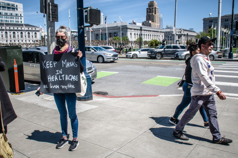 Two people walk past a lone demonstrator with sign: Keep Masks in Healthcare