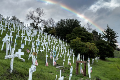 A rainbow arches above dozens of white crosses and symbols blanketing the hillside