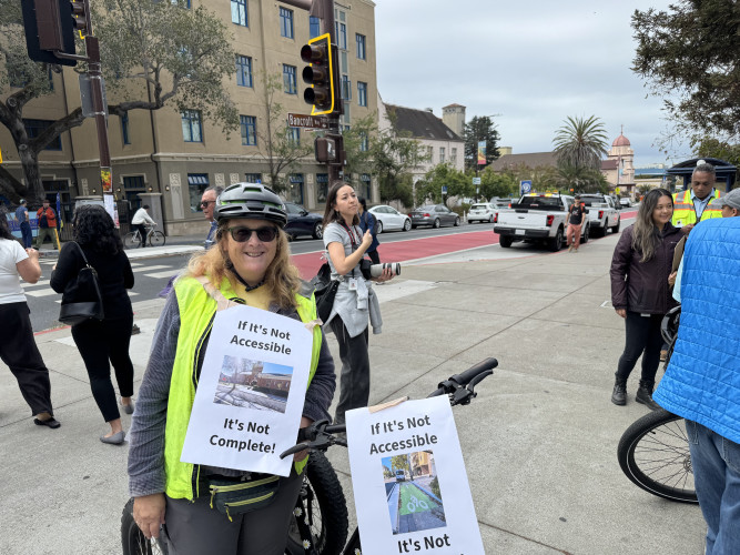 Helen and her adaptive cycle with daisies, wearing a sign that reads “If it’s not accessible, it’s not complete!”