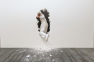 A white-clad figure passes through a torn opening in a pale surface, suspended between rupture and movement. 
