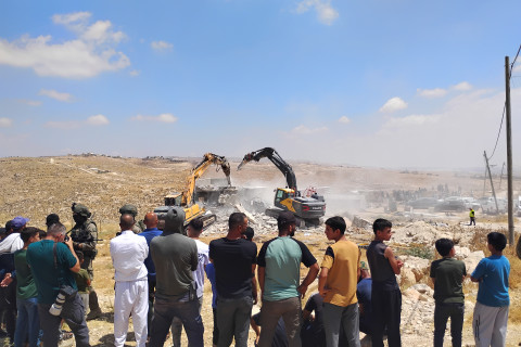 Foreground features a group of Palestinian villagers looking at construction equipment demolishing a home. 