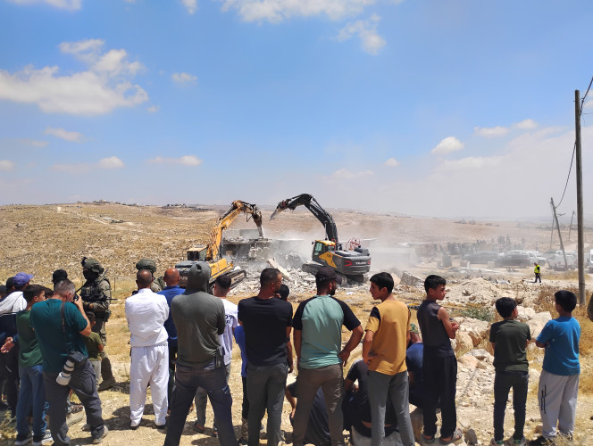 Foreground features a group of Palestinian villagers looking at construction equipment demolishing a home.